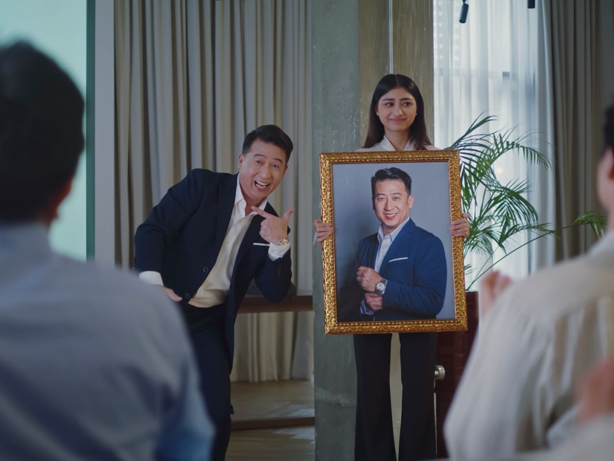 A grinning presenter in a suit points to a framed portrait of a man held up before a seated audience, from the GoTyme Bank campaign film.