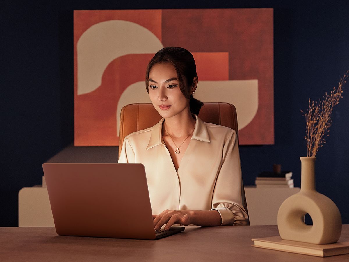 A woman working at a laptop in a warm-lit, elegant dark-blue office with abstract art behind her, from an Alcon campaign.