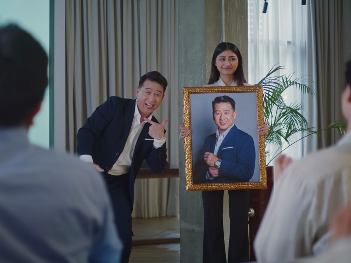 A grinning presenter in a suit points to a framed portrait of a man held up before a seated audience, from the GoTyme Bank campaign film.