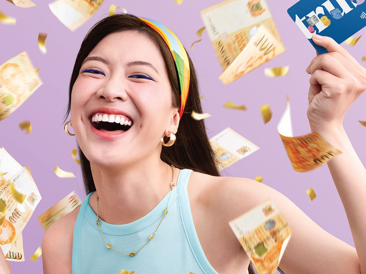 A woman laughing as Singapore dollar notes and gold confetti rain around her, holding up a Trust Bank cashback card.