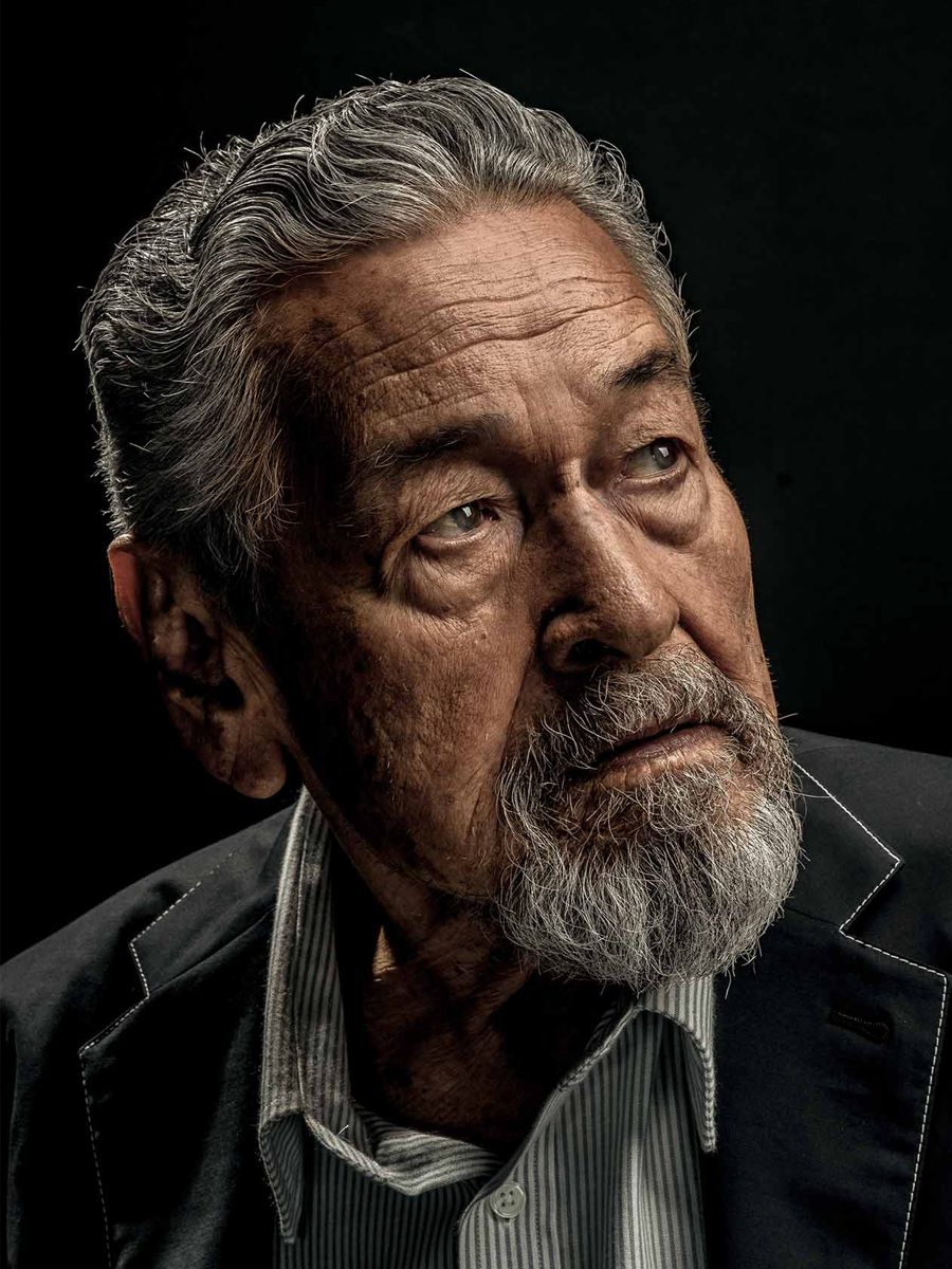 A dramatic, low-key editorial portrait of an older man against a black background.