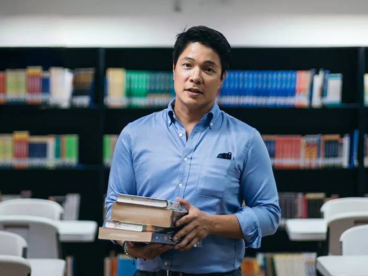A man in a blue shirt holds a stack of books in front of library shelves.